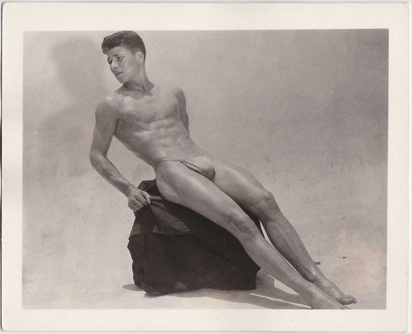 A handsome young man sits on a base, his long, lithe body doesn't fit in the frame. vintage gay photo AMG