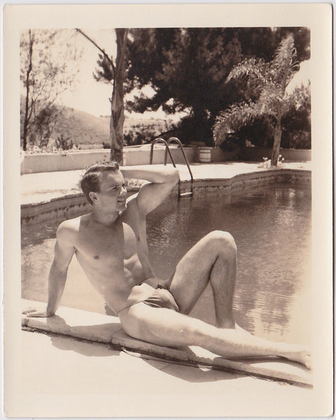 Handsome bodybuilder in posing strap reclines by the pool. Vintage physique photo
