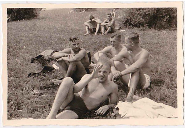 Several guys in their shorts, clothing scattered around them. The humpy man in the foreground thumbs his nose at the photographer.