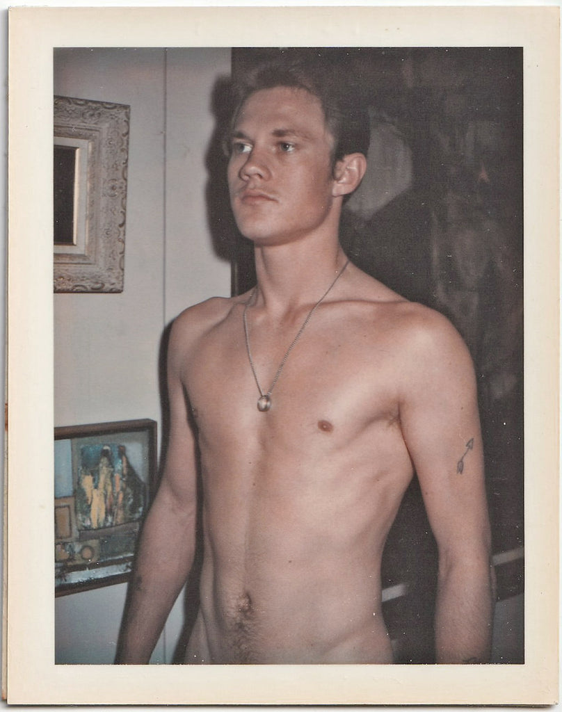 A vintage gay Polaroid of a male nude  standing next to a wall of paintings.