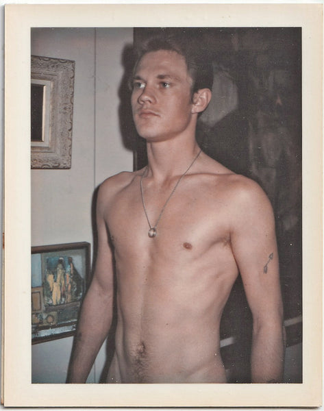 A vintage gay Polaroid of a male nude  standing next to a wall of paintings.