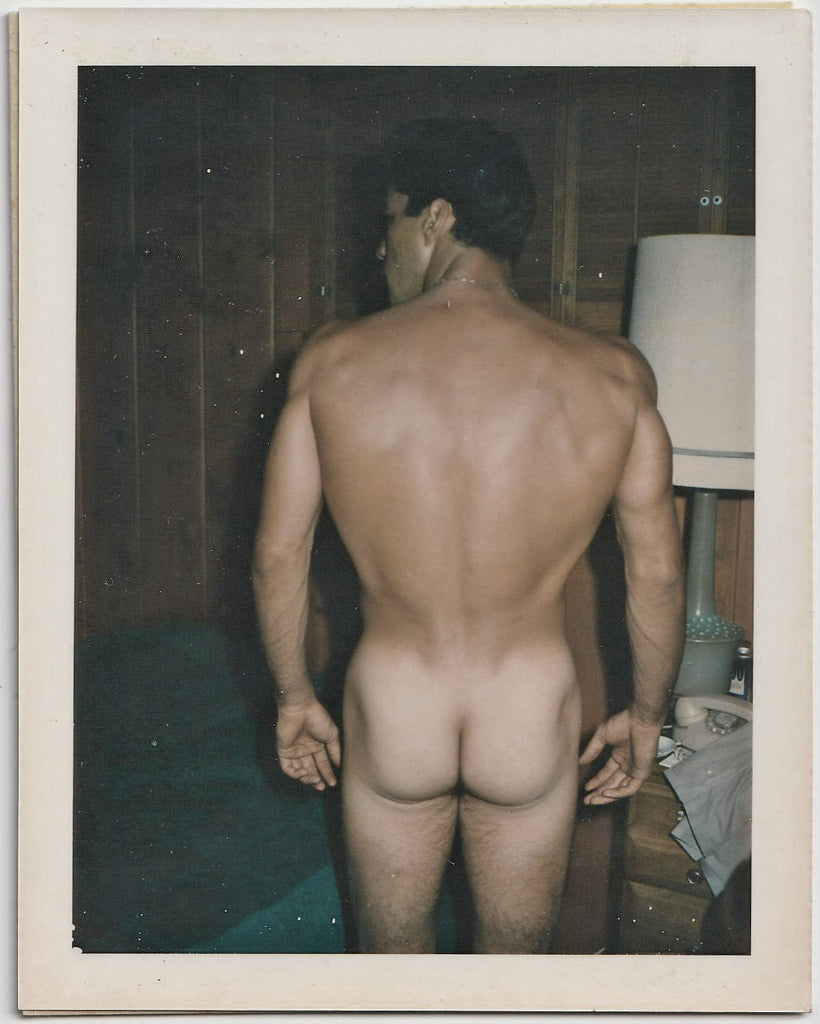 A vintage gay Polaroid Broad shoulders, slim waist, muscle butt male nude stands next to a cluttered bedside table. 