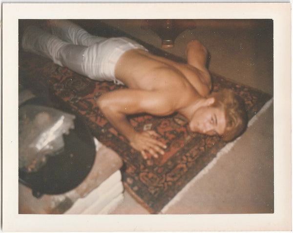 Vintage gay Polaroid Shirtless guy in white jeans looks up at the camera while doing a push-up on a Persian carpet.