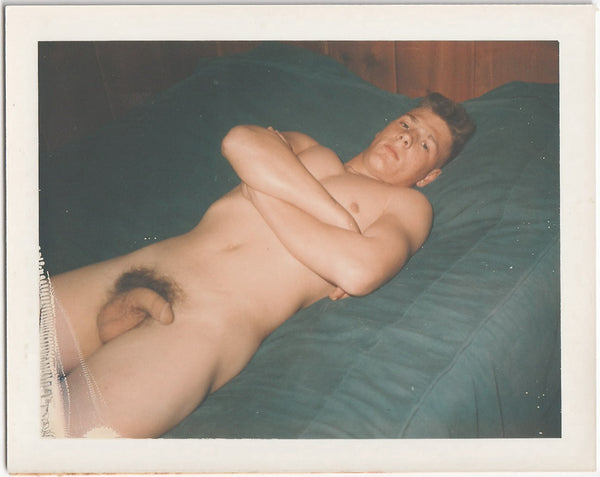 Vintage gay Polaroid Serious guy lies naked on the blue bedspread, arms crossed on his chest.