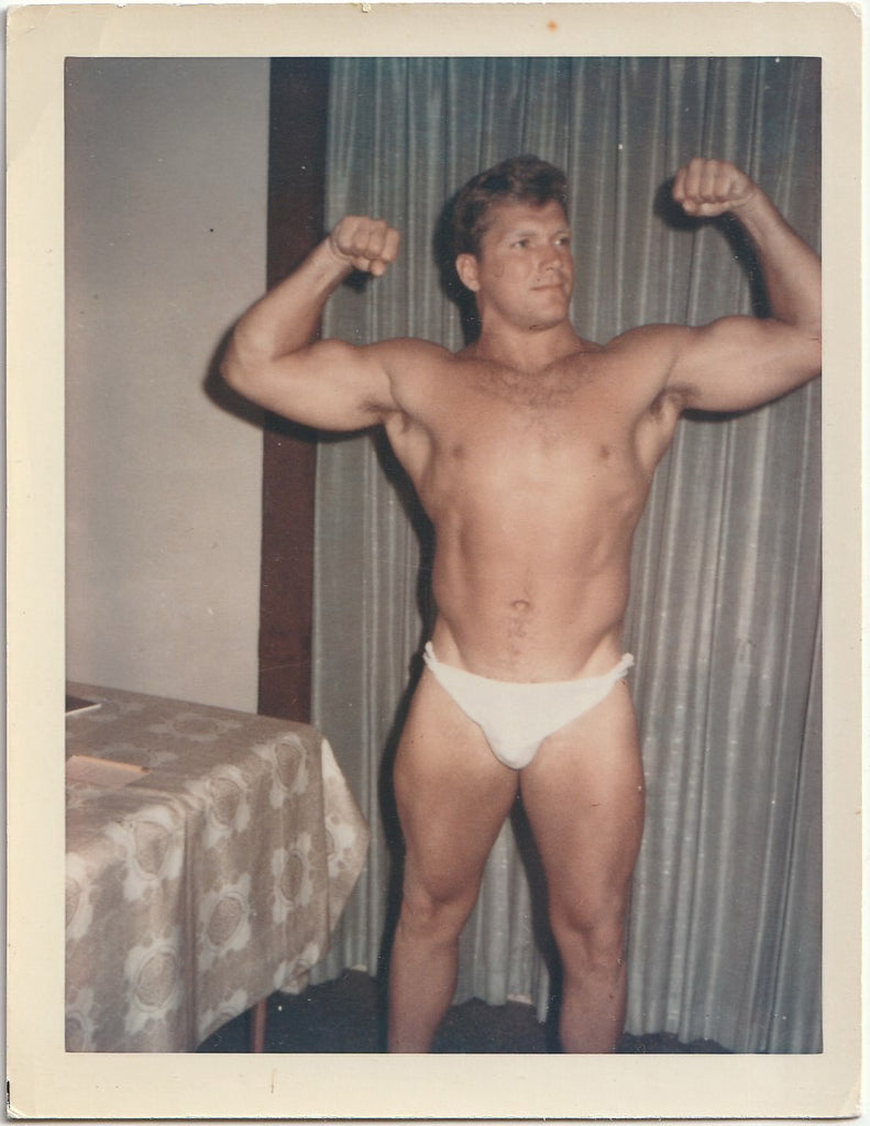 Vintage gay Polaroid, Handsome, muscular man flexes his biceps next to the dining room table.