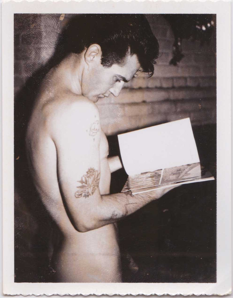 Vintage Polaroid Tattooed male nude standing before the fireplace looking at a gay porn magazine.