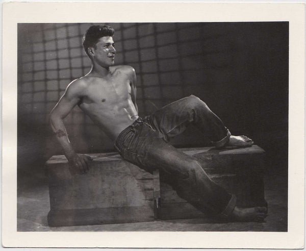 Against a projected net backdrop (courtesy of Athletic Model Guild) the shirtless, shoeless model reclines on a wooden case. Vintage gay photo AMG