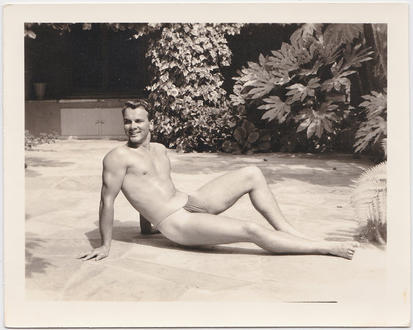 Vintage gay photo Handsome bodybuilder in posing strap lifts himself ever so slightly above the flagstone patio.