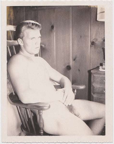 Male nude with a magnificent wave of blond hair seems a bit bored as he sits in a chair in knotty pine bedroom.