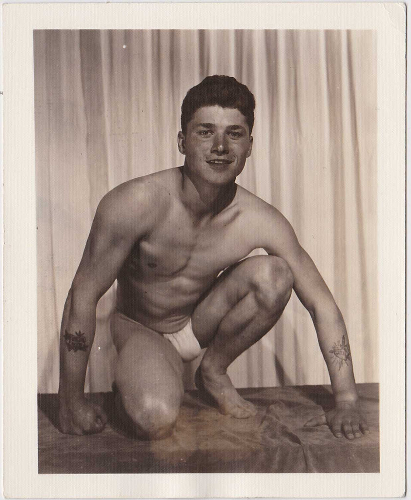 Vintage physique photo posing strap Handsome young guy with a tattoo on each forearm crouches