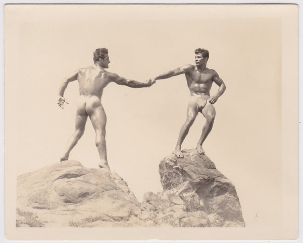 Great image of two nearly nude bodybuilders standing on boulders, reaching out to grasp each others hand
