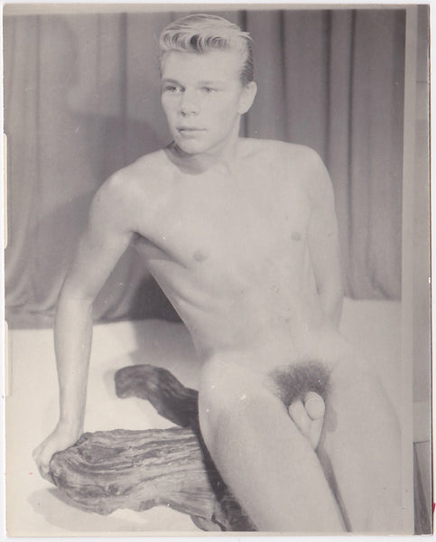Lithe blond male nude sits on a piece of driftwood.  Vintage physique photo