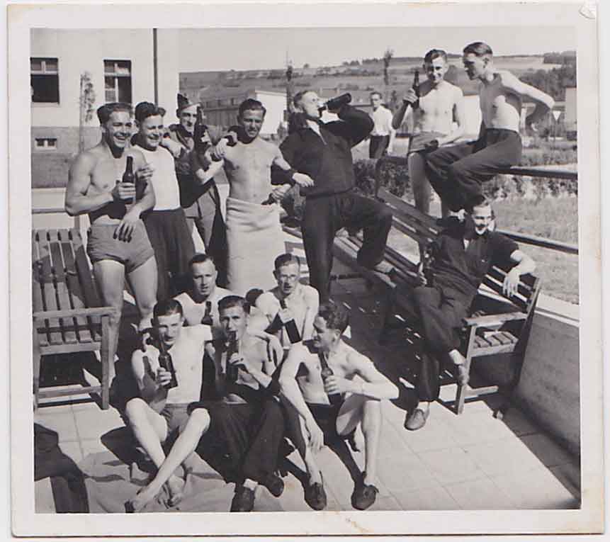 Group of men posing outdoors on a sunny day, some shirtless, others in swimsuits, drinking beer and laughing.