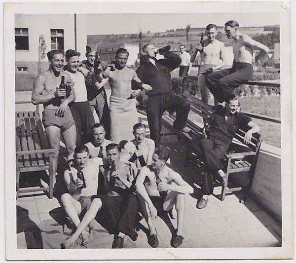 Group of men posing outdoors on a sunny day, some shirtless, others in swimsuits, drinking beer and laughing.