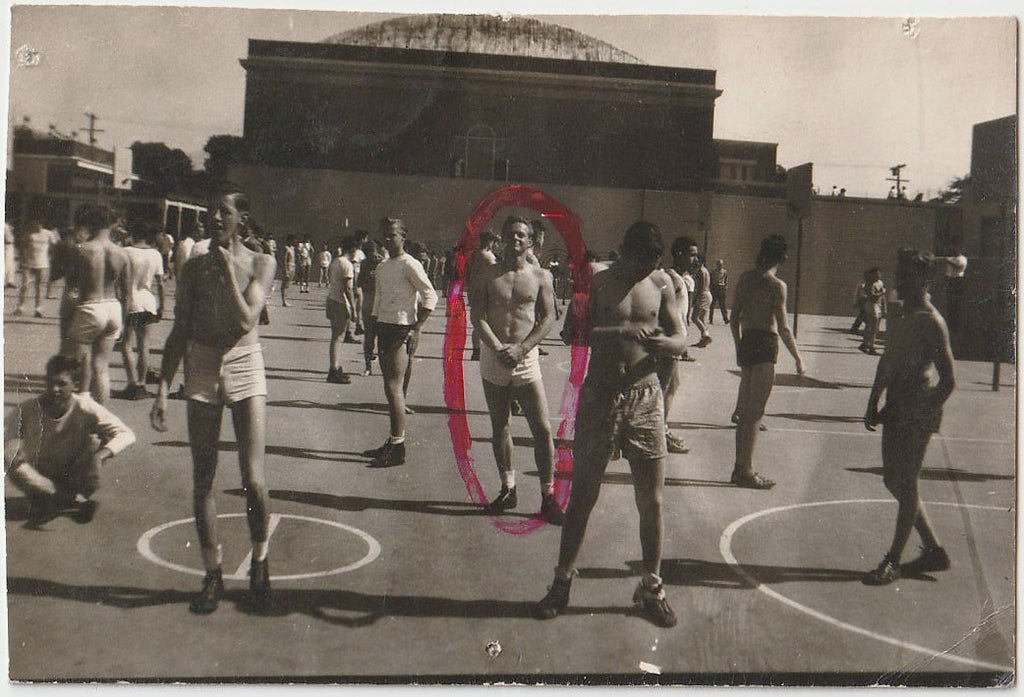 An urban court with dozens of shirtless guys hanging around but nobody seems to be playing anything. The stud in the center is circled in red ink.