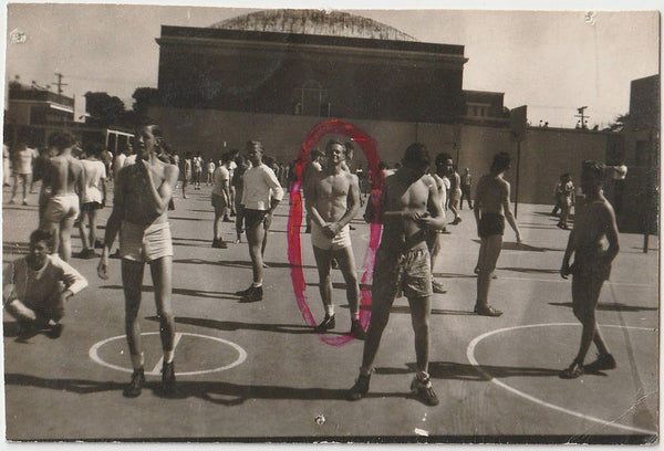 An urban court with dozens of shirtless guys hanging around but nobody seems to be playing anything. The stud in the center is circled in red ink.