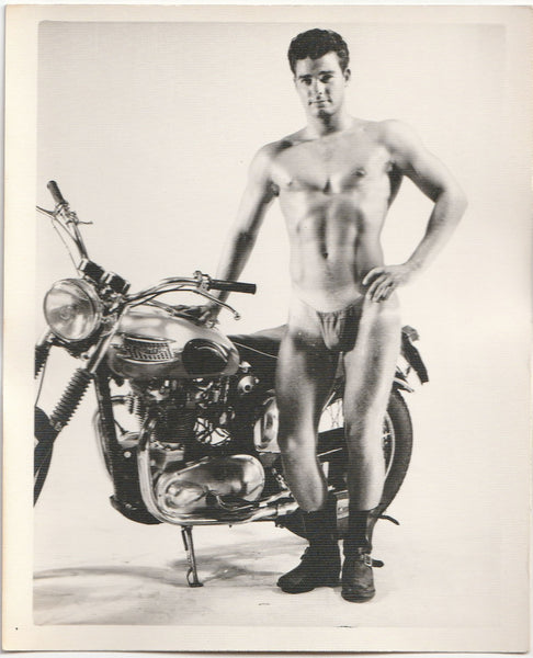 Handsome Jim Dillow, stripped down to his posing strap, proudly stands beside his Triumph motorcycle. By Kris Studio.