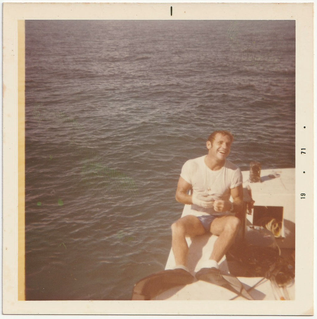 a handsome, hunky, laughing guy in swimsuit, t-shirt and flippers sits on a boat and removes his gloves. 1971
