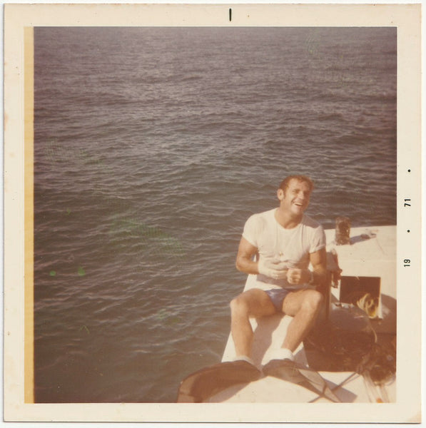 a handsome, hunky, laughing guy in swimsuit, t-shirt and flippers sits on a boat and removes his gloves. 1971
