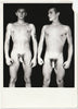 A black and white vintage gay physique photo showing two nude young men standing side by side against a dark background.