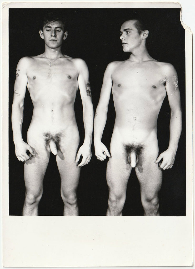A black and white vintage gay physique photo showing two nude young men standing side by side against a dark background.