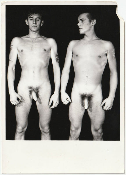A black and white vintage gay physique photo showing two nude young men standing side by side against a dark background.