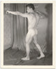 A black and white photograph of a nude male model Bob Raffael posing in profile against a draped studio backdrop by Kris Studio, Chuck Renslow.
