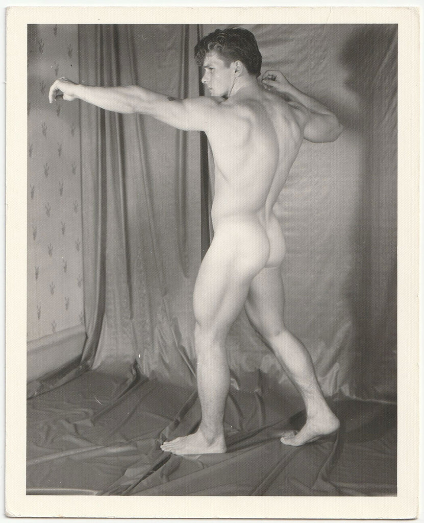 A black and white photograph of a nude male model Bob Raffael posing in profile against a draped studio backdrop by Kris Studio, Chuck Renslow.