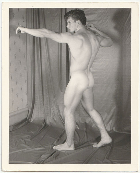 A black and white photograph of a nude male model Bob Raffael posing in profile against a draped studio backdrop by Kris Studio, Chuck Renslow.
