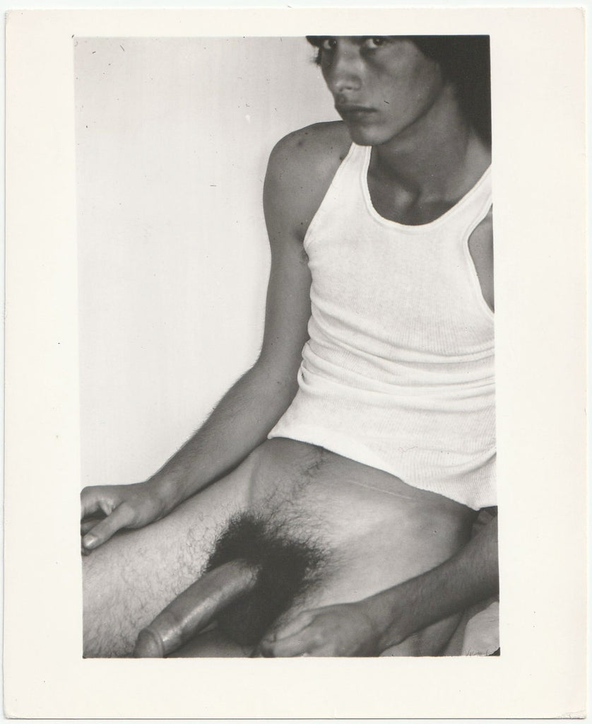 Black and white gay photo of a man in a white tank top, shown seated with his large penis exposed.