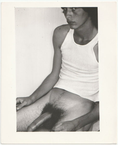 Black and white gay photo of a man in a white tank top, shown seated with his large penis exposed.