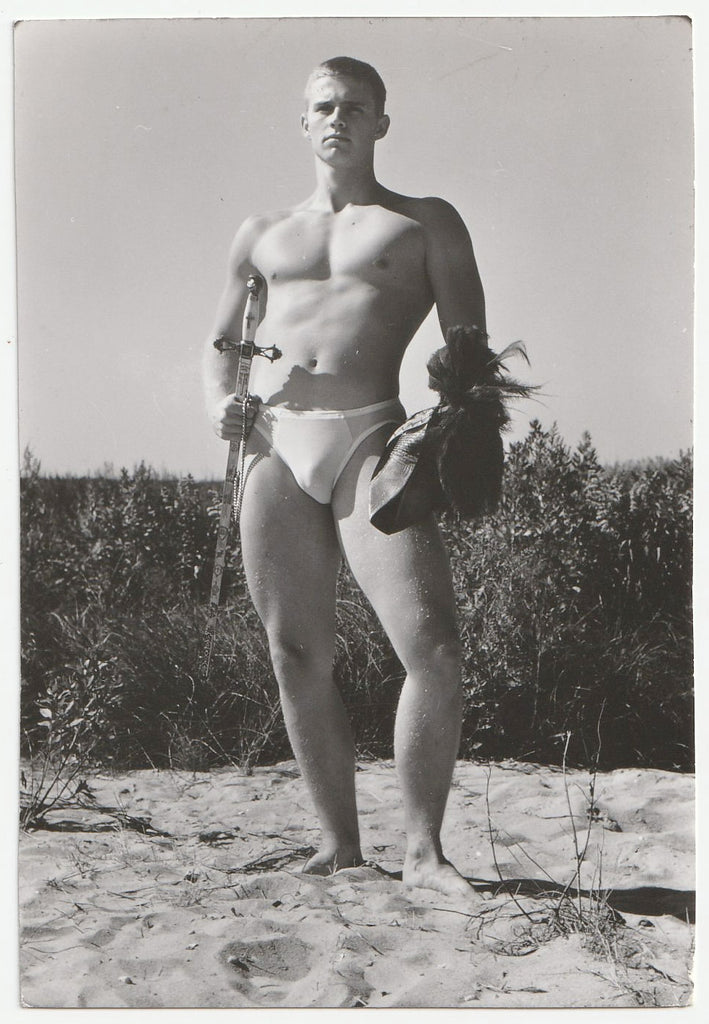 Vintage gay physique photo handsome hunk holds helmut and sword while standing on the beach.