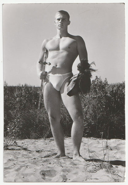 Vintage gay physique photo handsome hunk holds helmut and sword while standing on the beach.