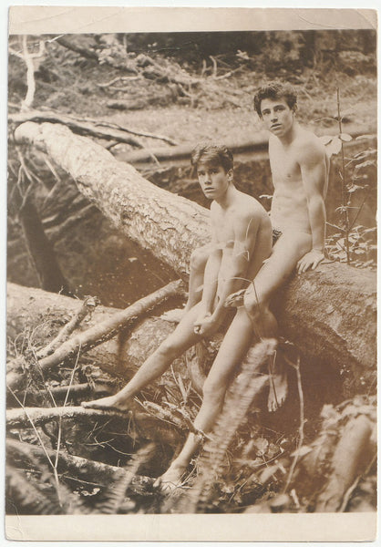 Two naked guys sitting on a fallen tree. One sits between the legs of the other.