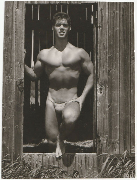 Handsome bodybuilder in posing strap stands in the opening of a barn window.