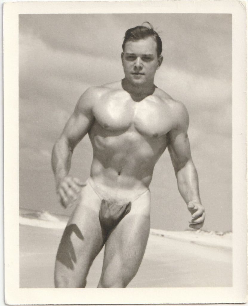 Cute muscle guy with nice tan lines jogs on an empty beach wearing only his posing strap. Vintage physique photo.