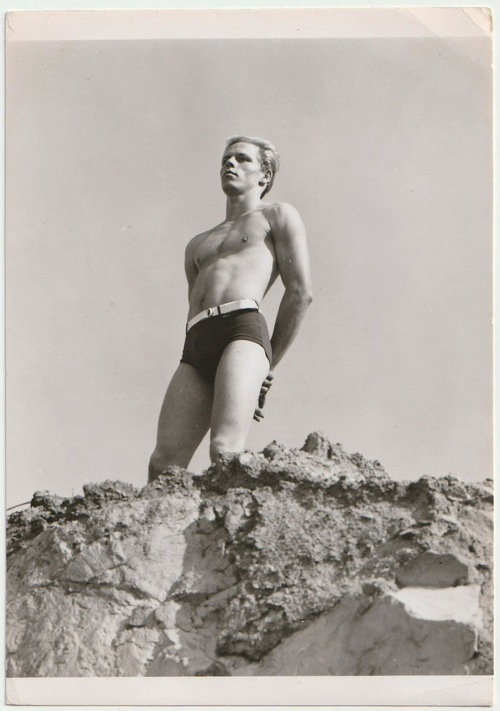 Handsome blond guy wearing fashionable swim trunks stands near the edge of a sandy cliff.