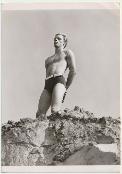 Handsome blond guy wearing fashionable swim trunks stands near the edge of a sandy cliff.