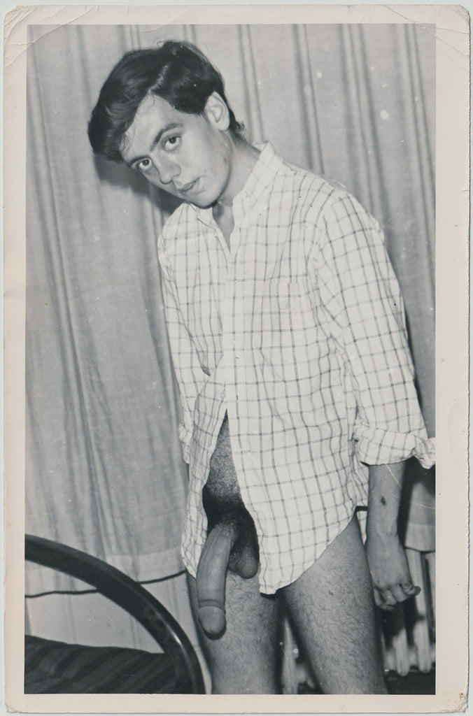 A vintage black and white photo of a young man wearing only a shirt and standing beside his bed.