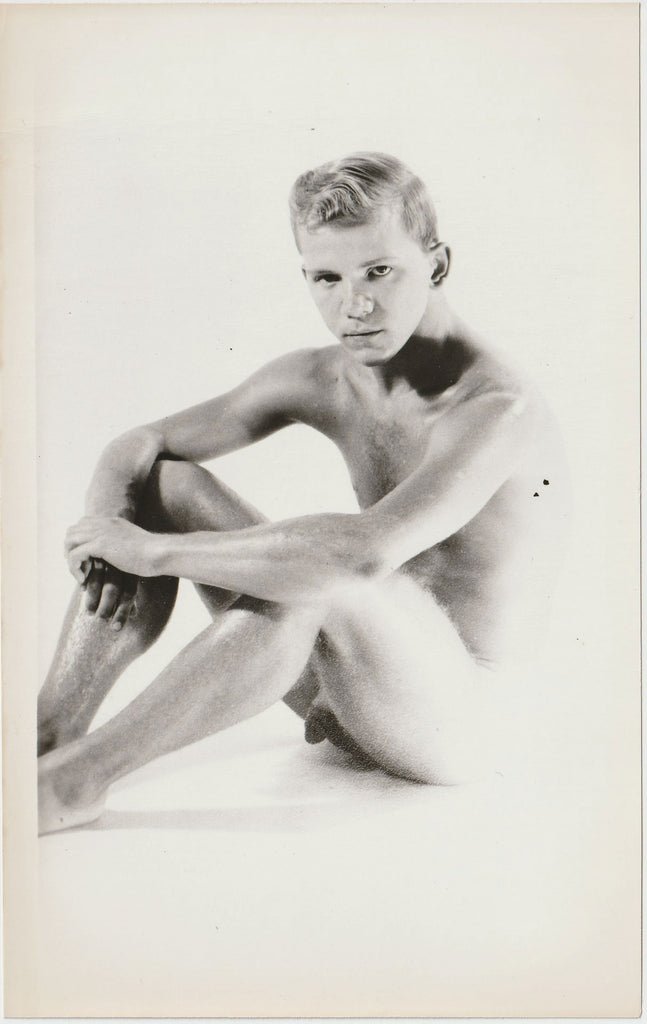 A black and white artistic studio photograph of a seated nude male figure.