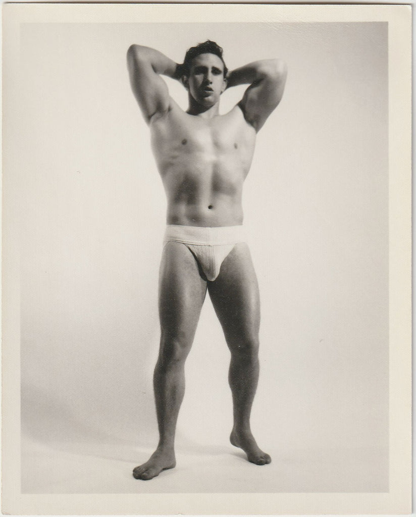 A black and white vintage physique photograph of a muscular man posing in a jockstrap by Bruce of LA.