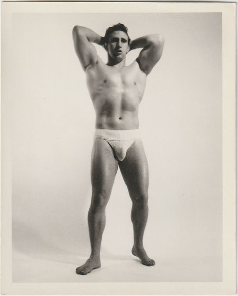 A black and white vintage physique photograph of a muscular man posing in a jockstrap by Bruce of LA.