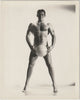 A black and white studio physique photograph of a muscular man standing barefoot and holding a ball between his legs, by Bruce of LA,