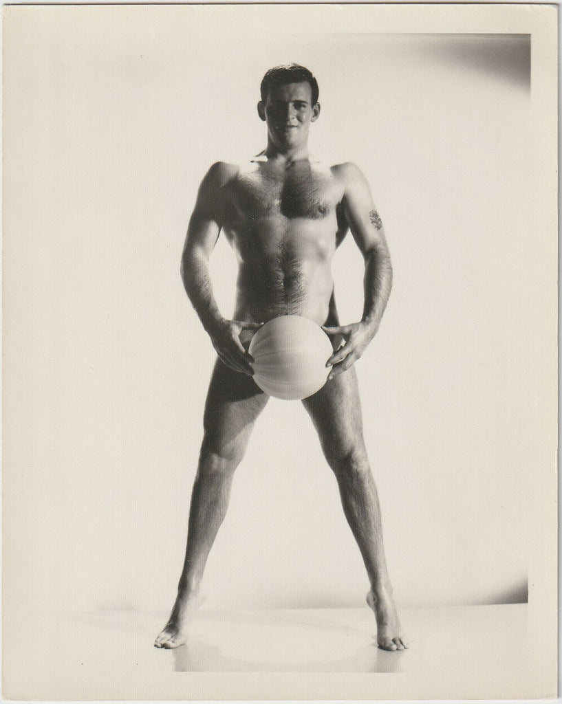 A black and white studio physique photograph of a muscular man standing barefoot and holding a ball between his legs, by Bruce of LA,