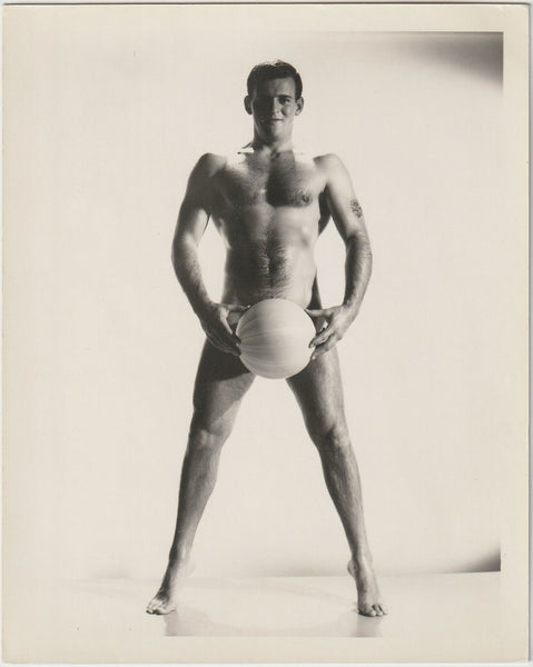 A black and white studio physique photograph of a muscular man standing barefoot and holding a ball between his legs, by Bruce of LA,