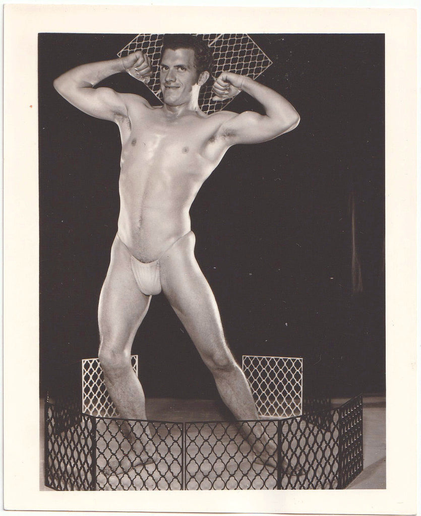 A black and white vintage gay physique photograph of a muscular male model in a posing strap flexing his biceps.