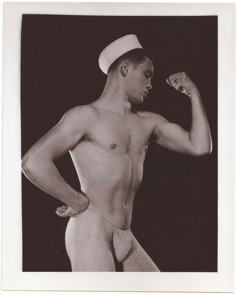 A vintage black and white physique photograph print showing a muscular man posing in a sailor hat posing strap.