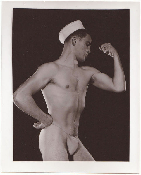A vintage black and white physique photograph print showing a muscular man posing in a sailor hat posing strap.