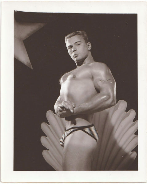 A vintage black and white gay physique photo of a muscular male bodybuilder posing in striped bikini-style underwear.