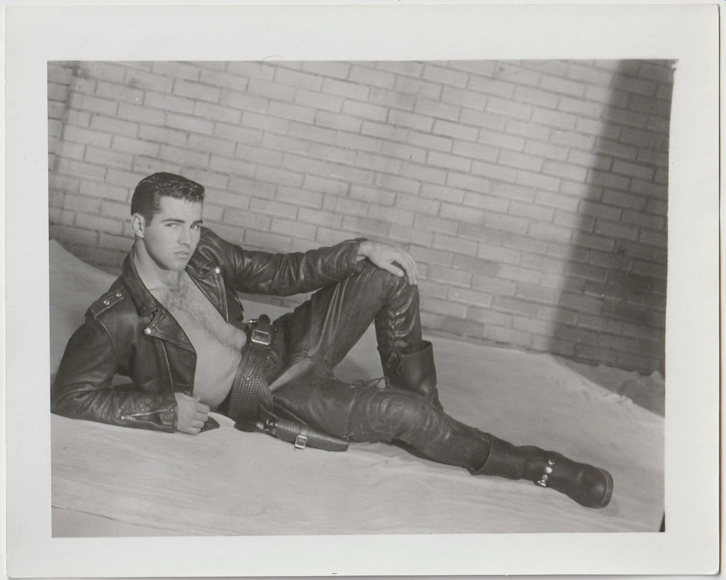 A black and white vintage gay photograph featuring a man in a leather jacket and leather pants reclining in front of a wall.